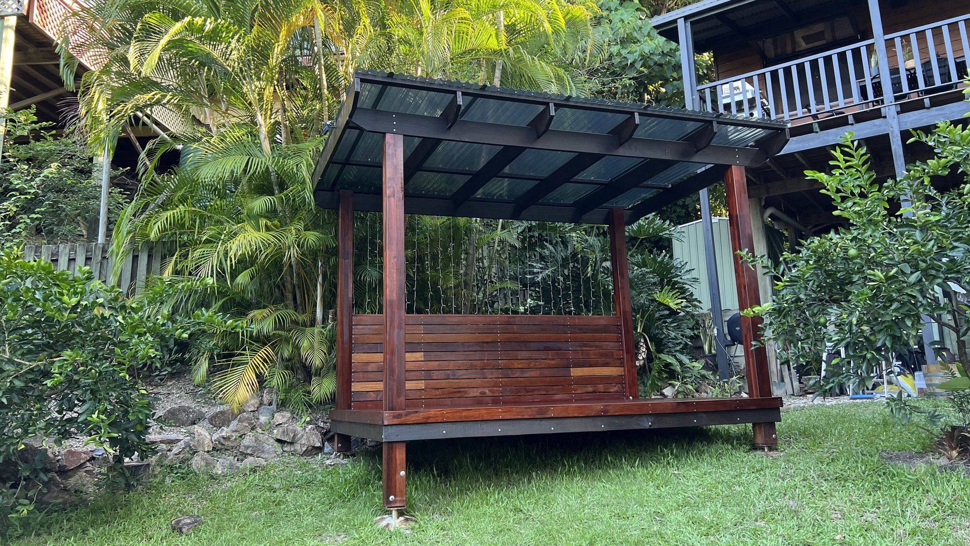 A hardwood gazebo with a corrugated roof standing in a garden below two raised timber homes