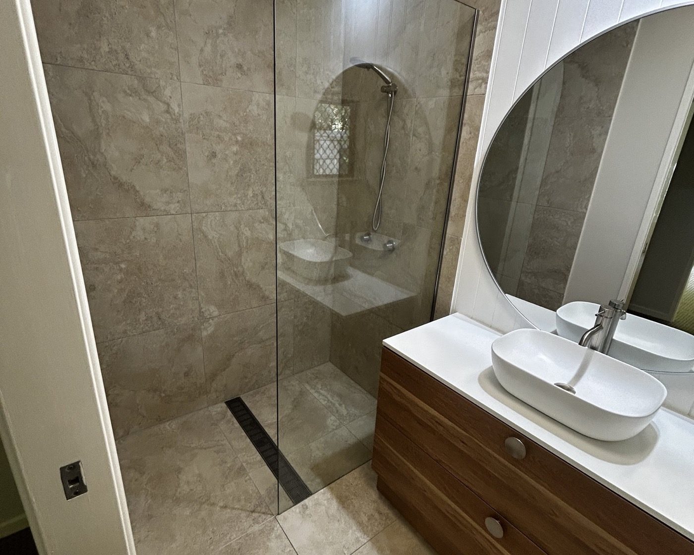 A renovated bathroom with a timber vanity, round mirror and glass shower screen