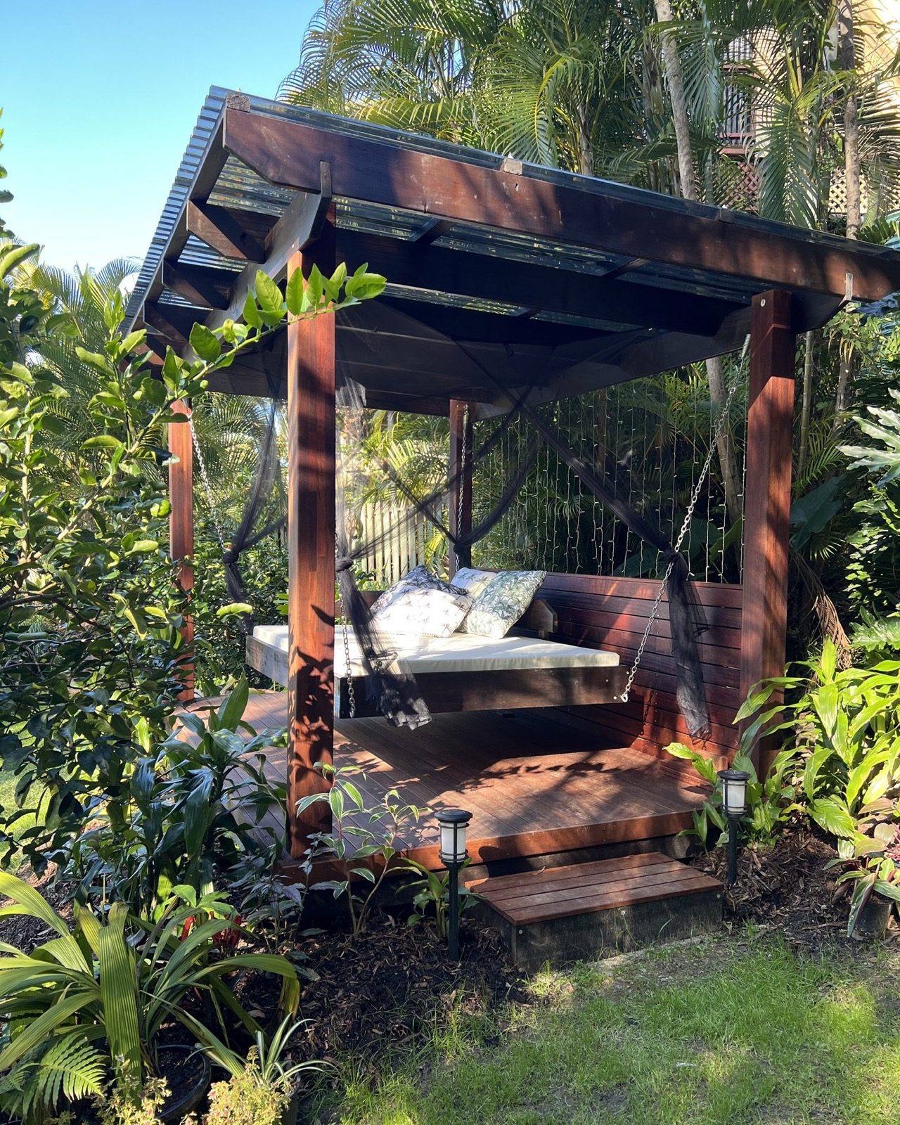 A hardwood pergola with a slatted screen and a hanging daybed in a palm garden