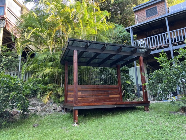 A hardwood gazebo with a corrugated roof standing on a lawn below two raised timber homes